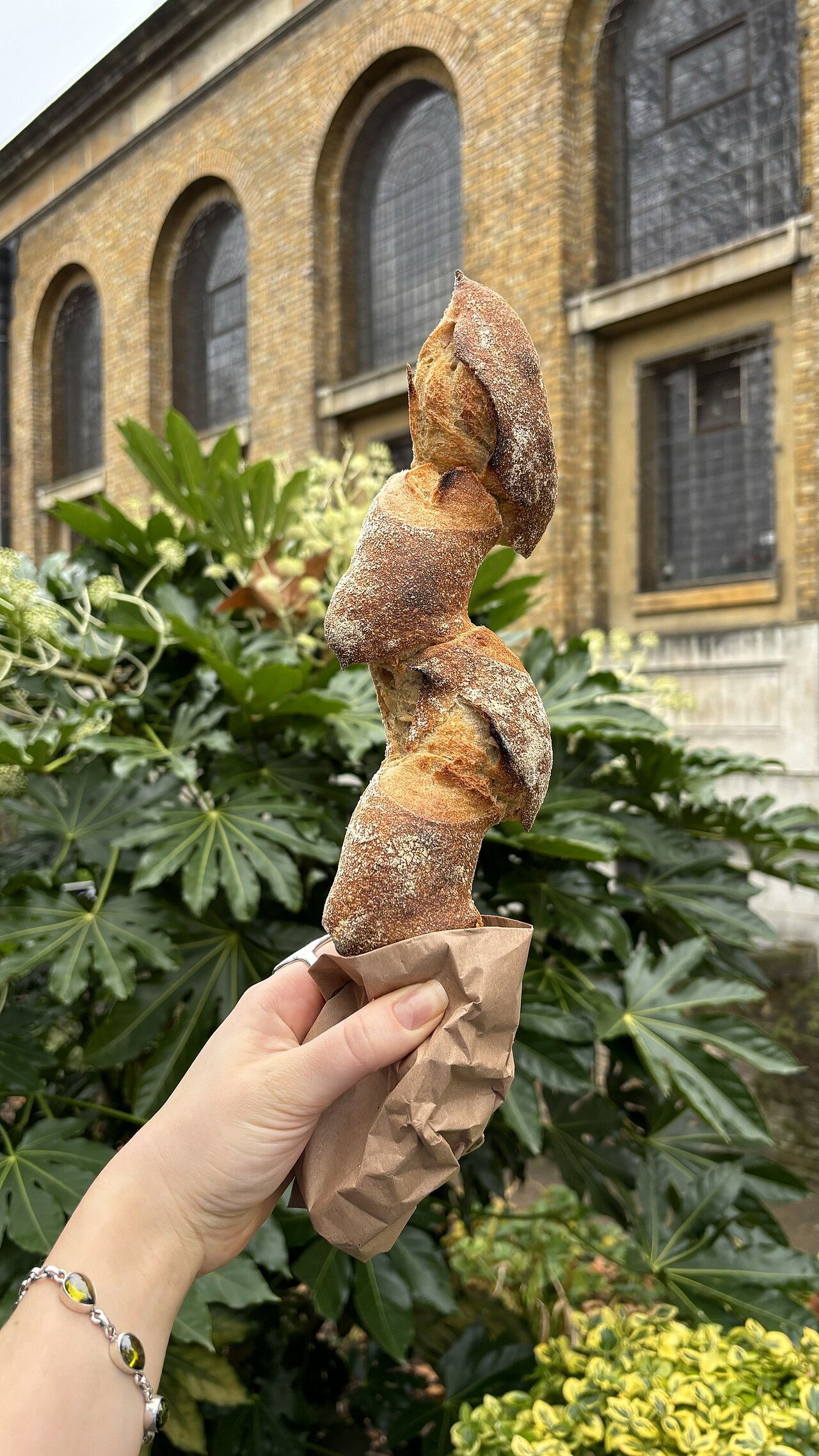 Hand holding golden-brown pain d'épi bread wrapped in tan paper outdoors