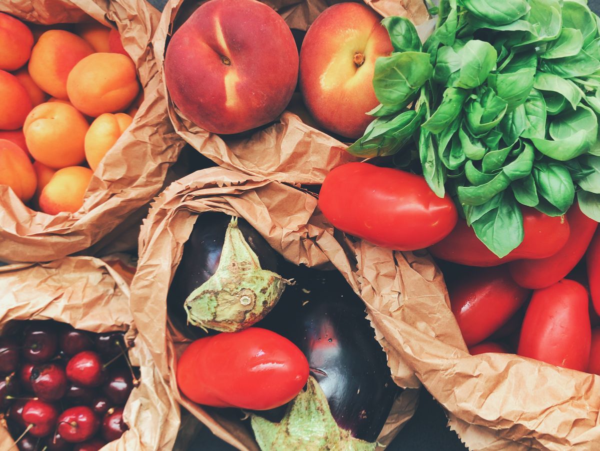 Overhead view of paper bags filled with apricots, peaches, basil, tomatoes, eggplants, and cherries