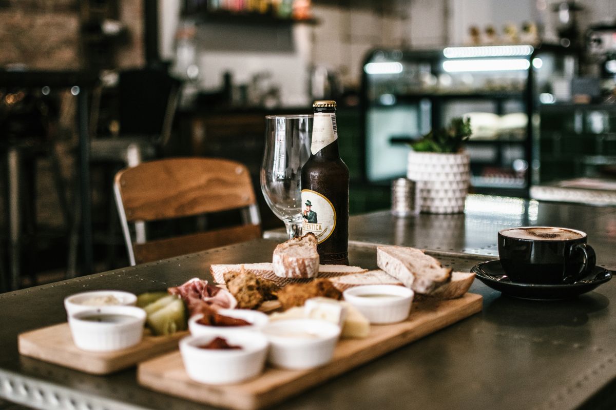 Wooden boards with bread, cured ham, cheese, dips, a beer bottle, and a foam-topped coffee cup