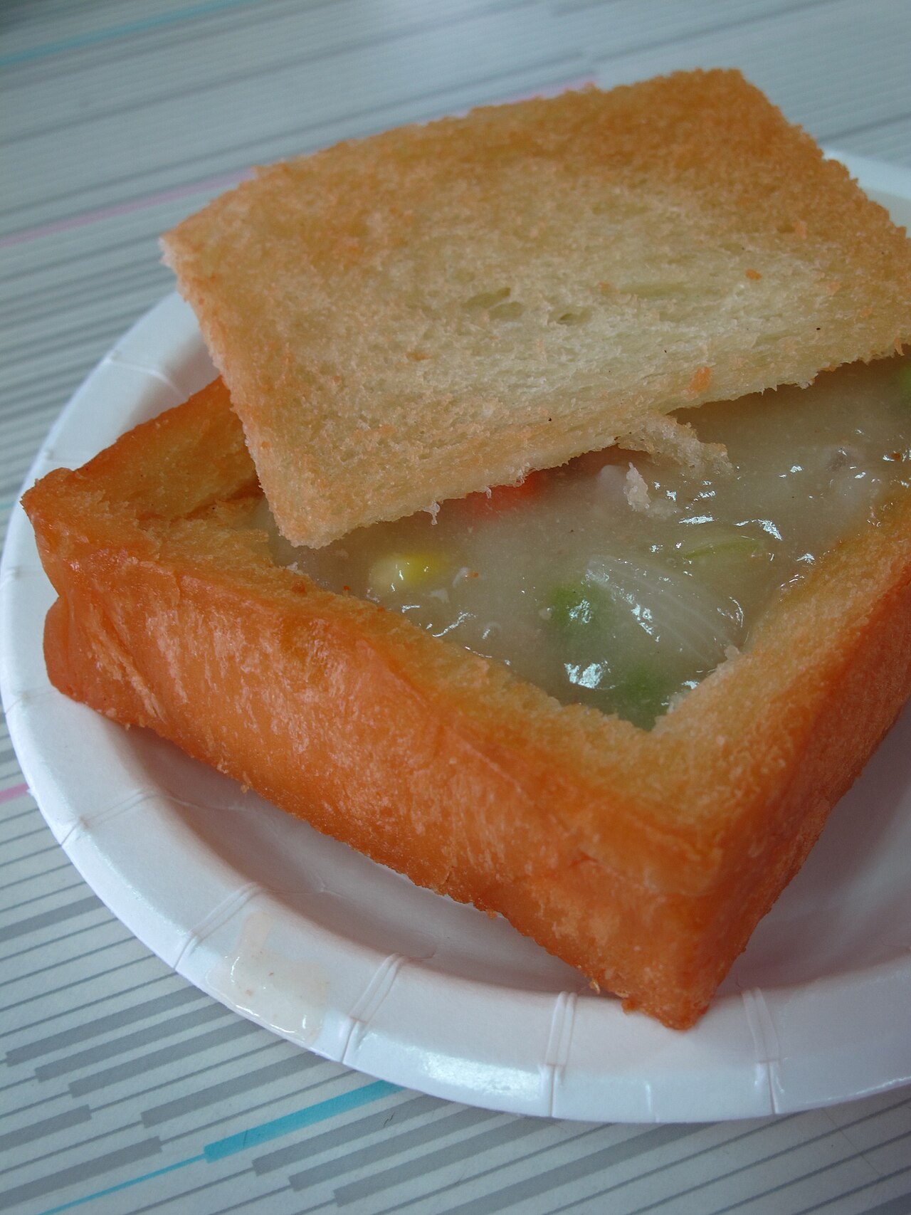 Square hollowed fried bread filled with creamy white stew and vegetables on a paper plate