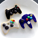 Three iced sugar cookies shaped like video game controllers in black, purple, and light gray