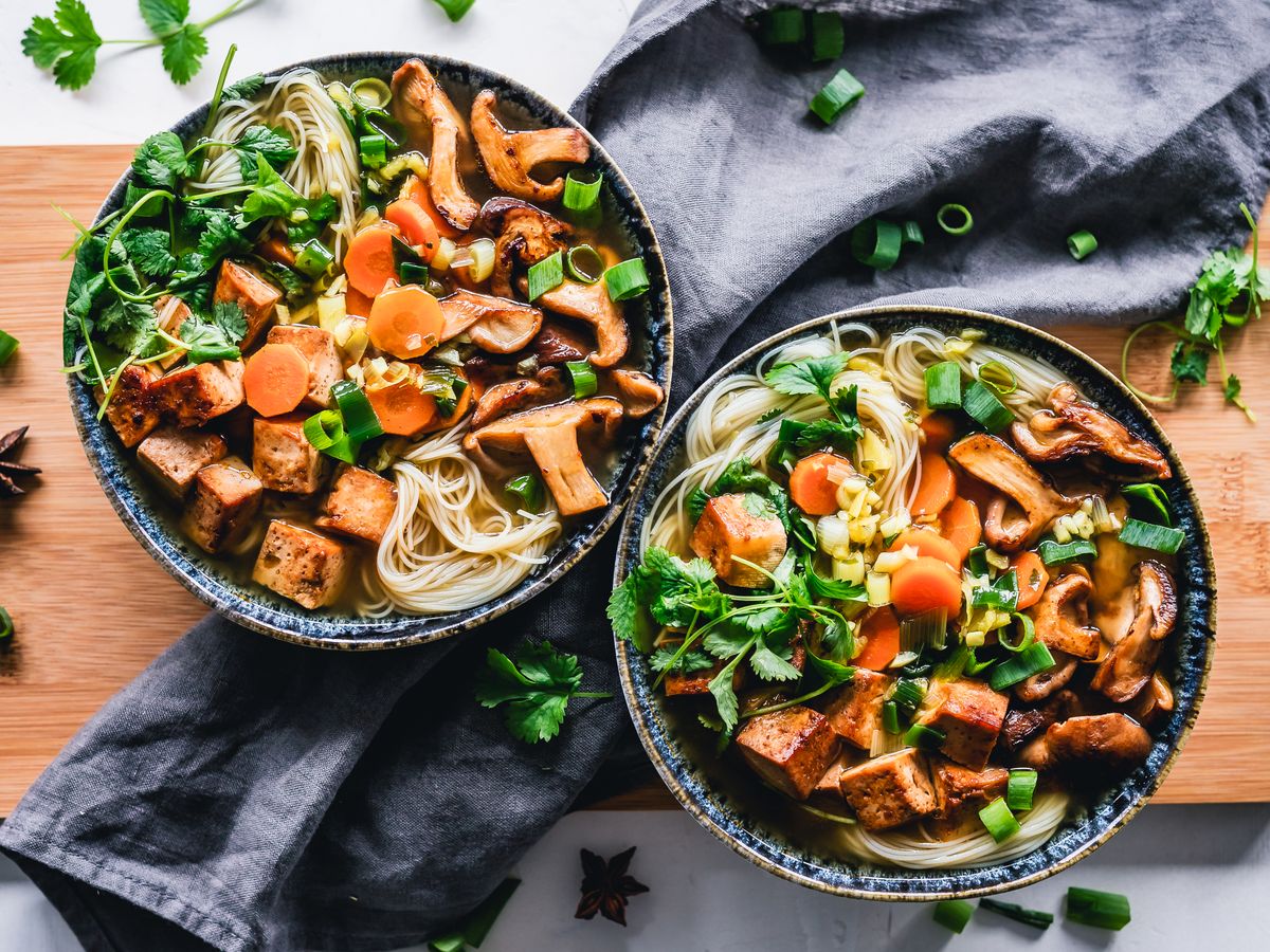 Two bowls of noodle soup with seared tofu, mushrooms, carrots, and cilantro on a wooden board