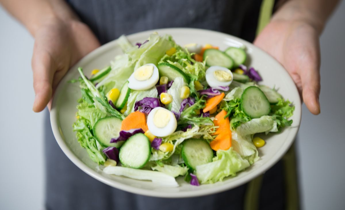Hands holding a gray plate of mixed salad with cucumber, carrots, cabbage, corn, and quail eggs