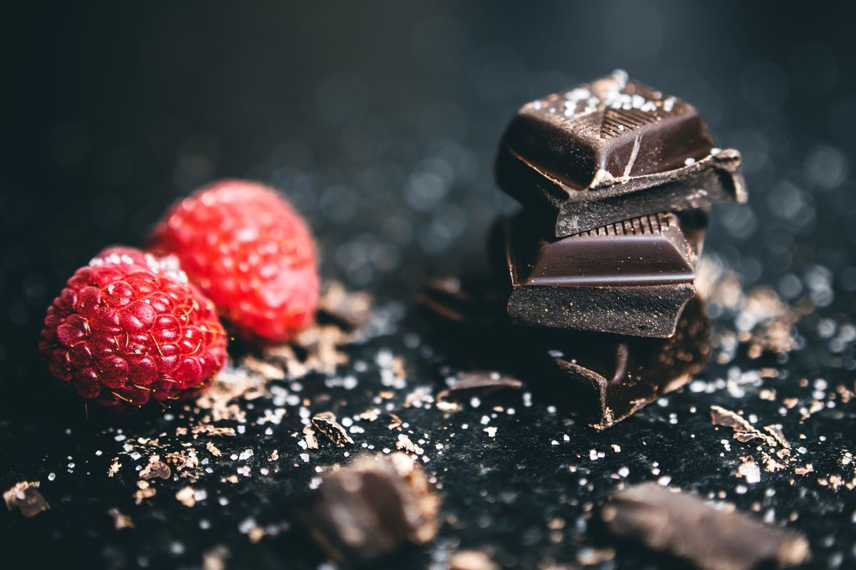 Stacked dark chocolate squares with coarse salt beside two fresh red raspberries on dark stone