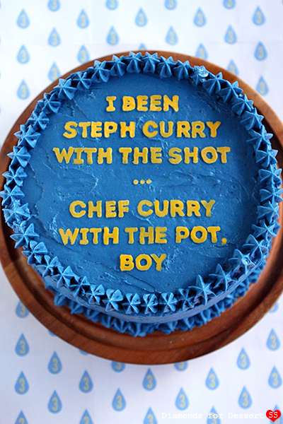 Top-down view of blue-frosted round cake with piped star borders and yellow block lettering