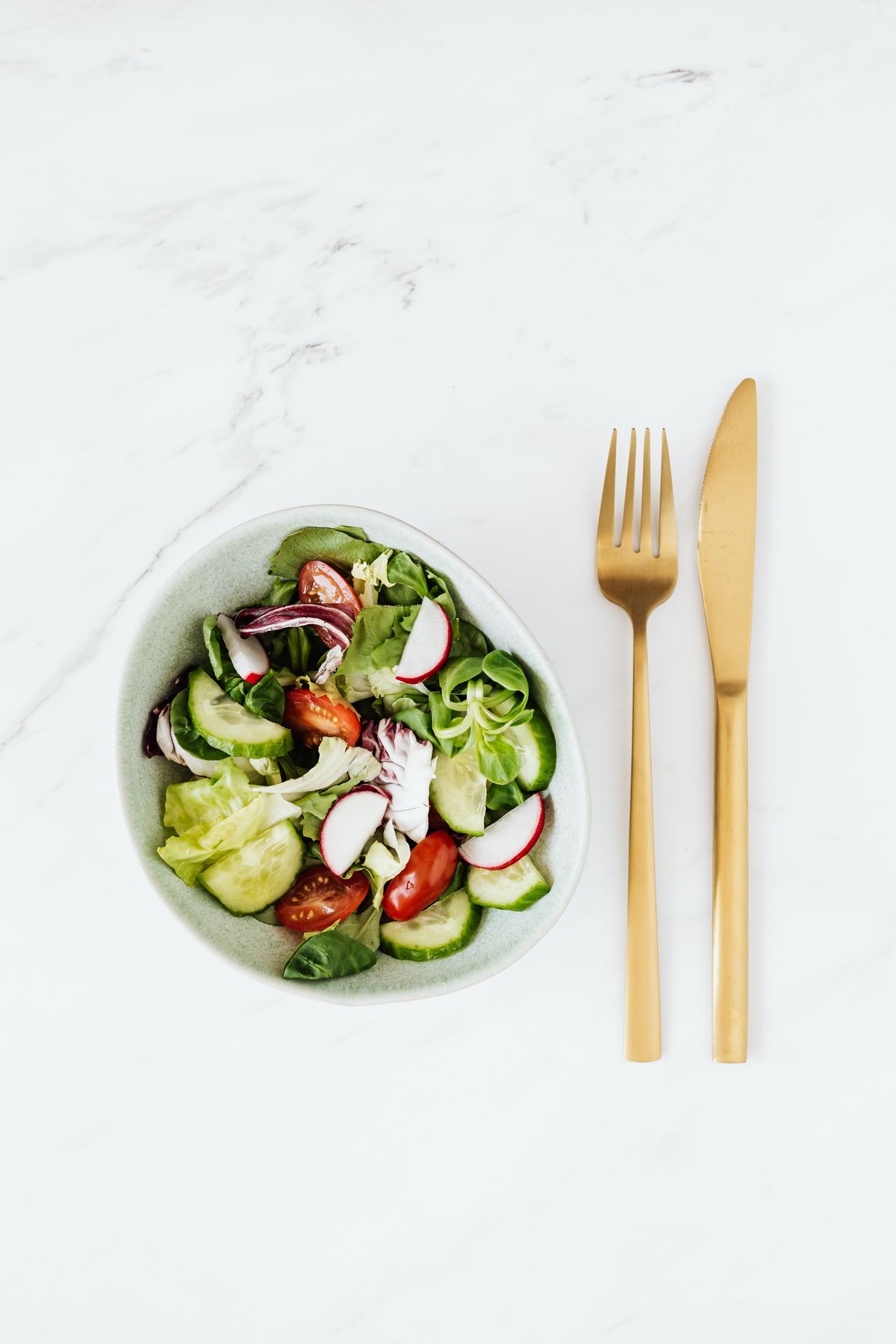 Top-down garden salad in a gray bowl beside gold cutlery on white marble
