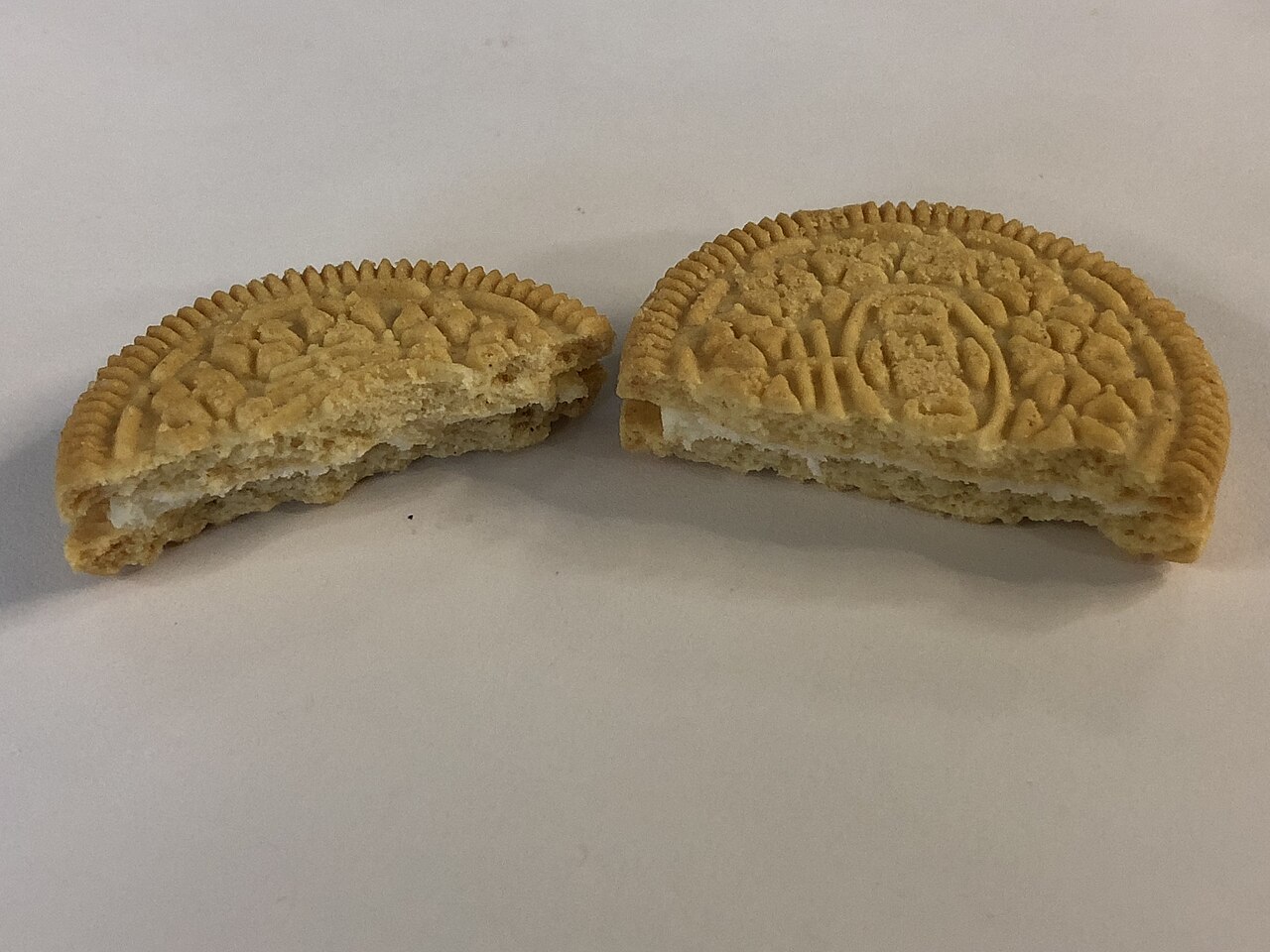 Two broken pieces of a golden sandwich cookie with white cream filling on a light grey surface