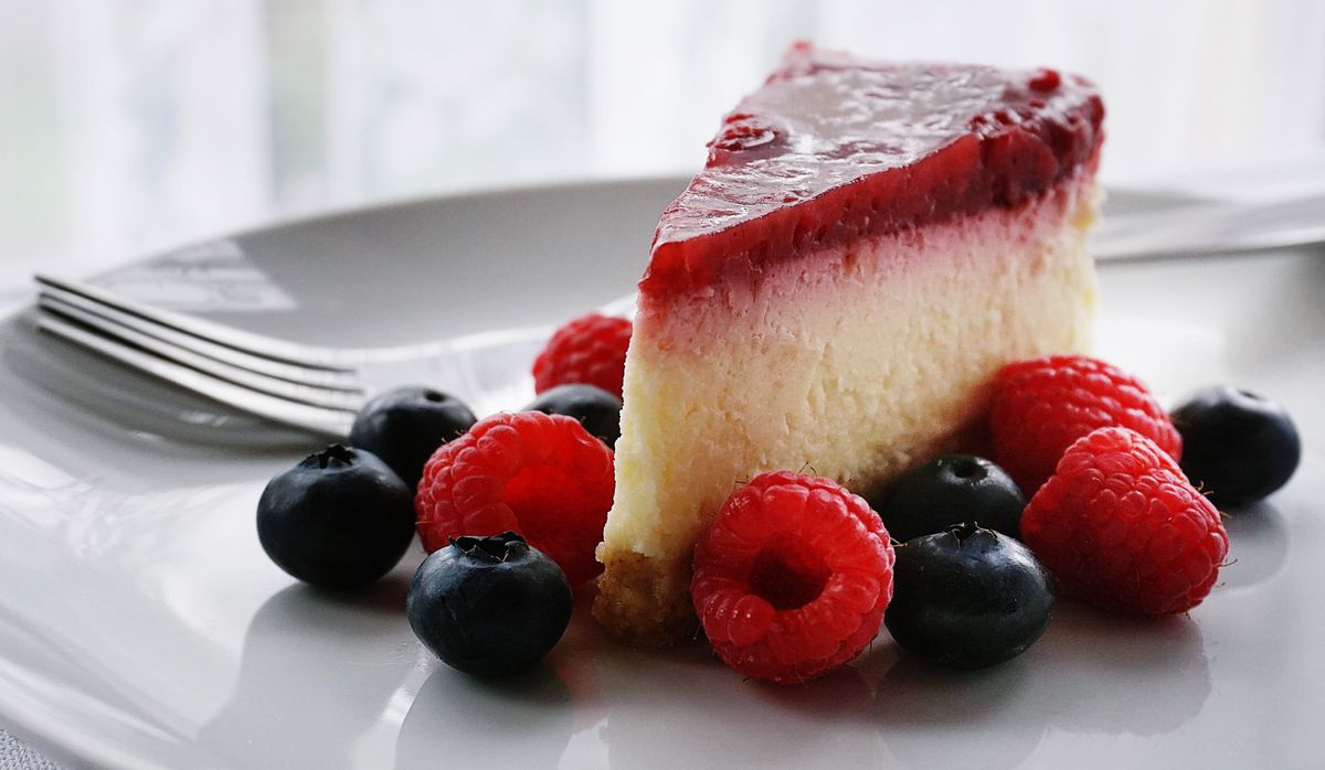 Slice of berry cheesecake with red glaze topped with blueberries and raspberries on a white plate