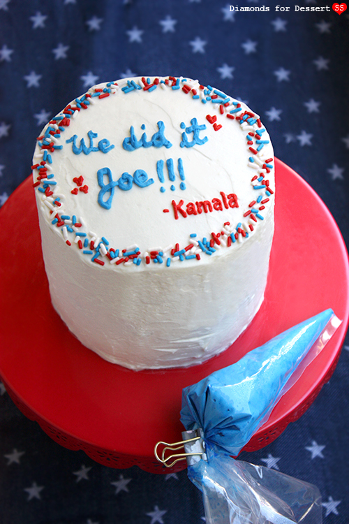 Tall white-frosted cake on a red stand reading We did it Joe and Kamala in blue and red icing with sprinkles