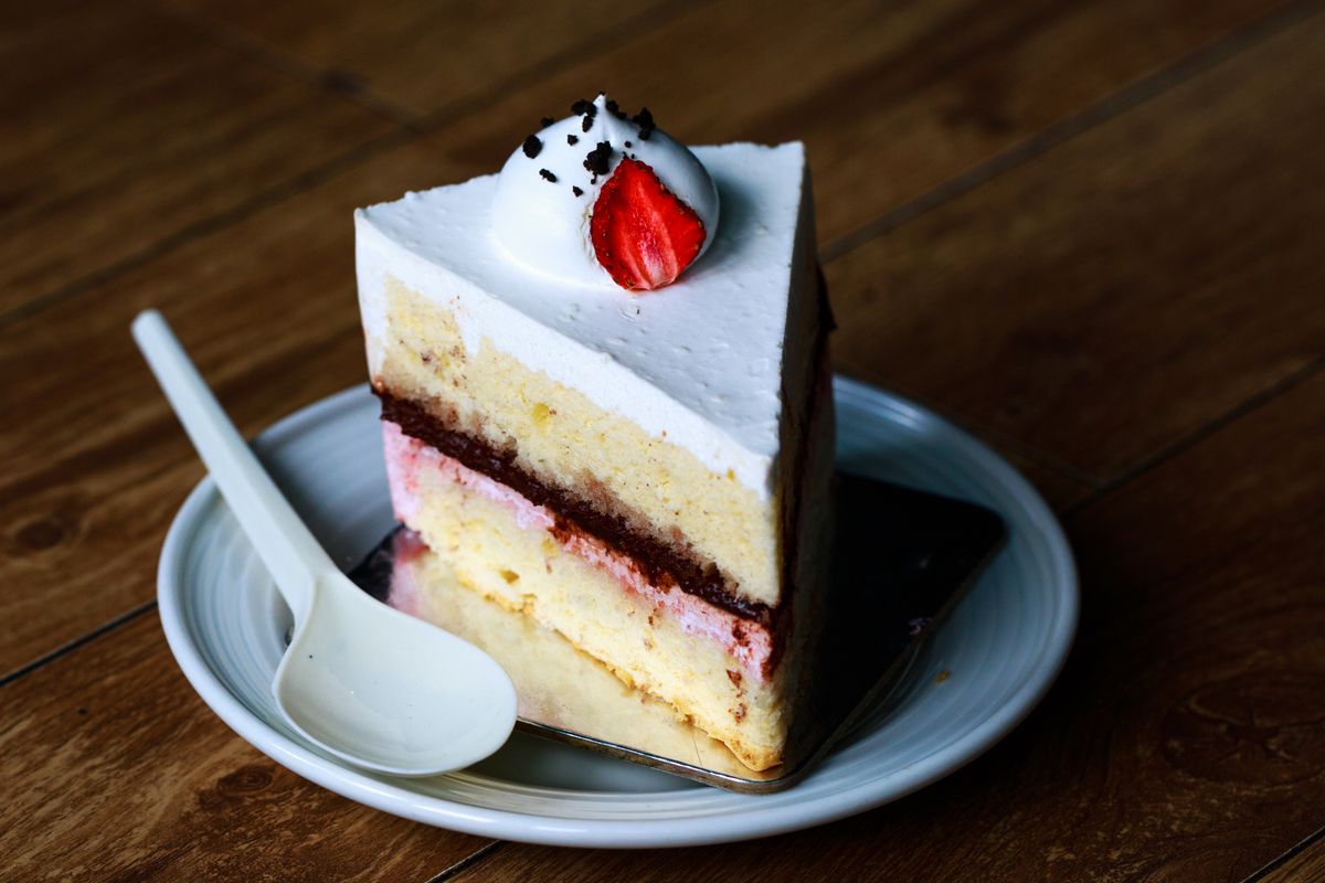 Triangular cake slice with sponge layers, pink cream, red jam, white frosting, and a strawberry