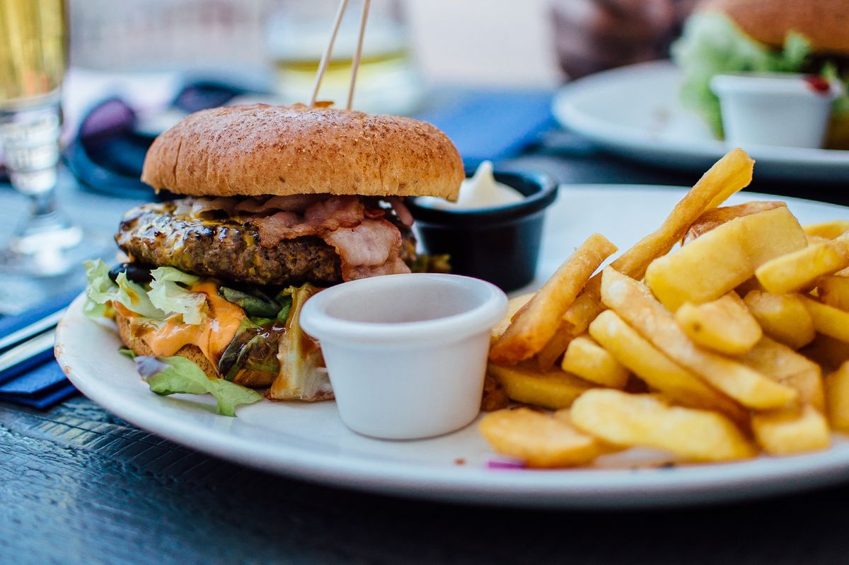Bacon cheeseburger with thick fries on a white plate beside small sauce cups