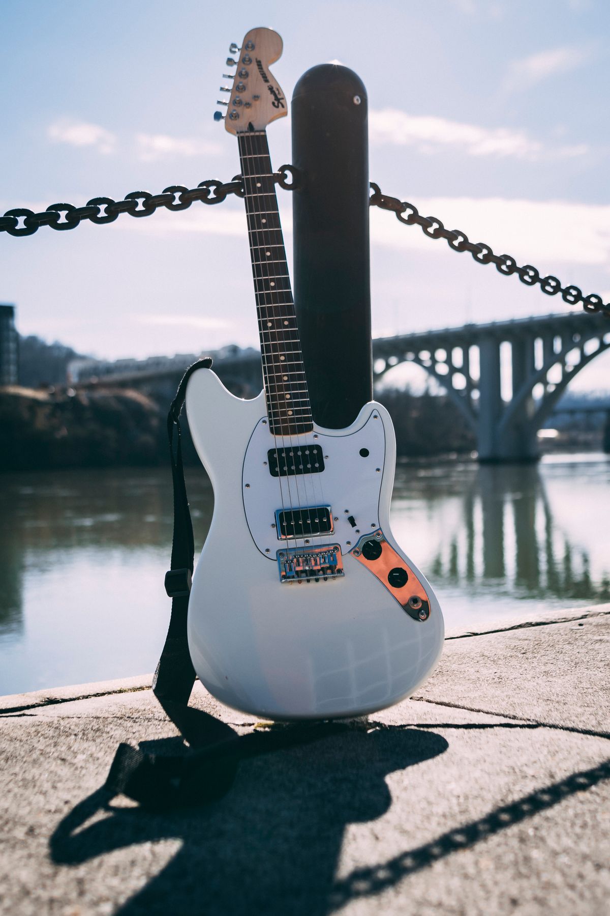 White Squier Mustang electric guitar leaning against a metal post on a waterfront walkway