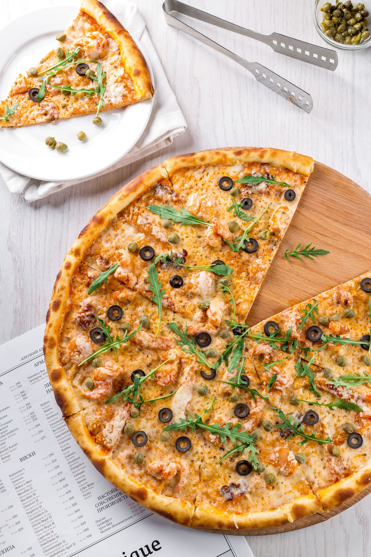 Thin-crust pizza with shrimp, olives, capers, and arugula on a wooden board