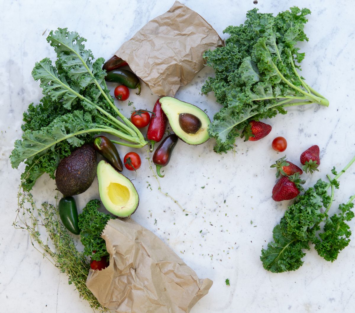 Flat lay of fresh kale, avocado, tomatoes, peppers, and strawberries on white marble
