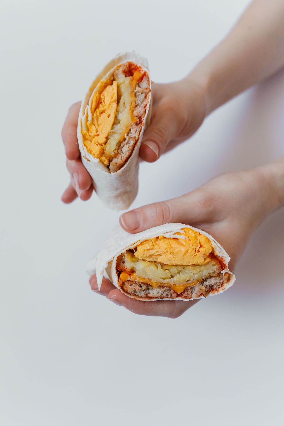 Hands holding two halves of a breakfast burrito showing scrambled eggs, potatoes, and sausage