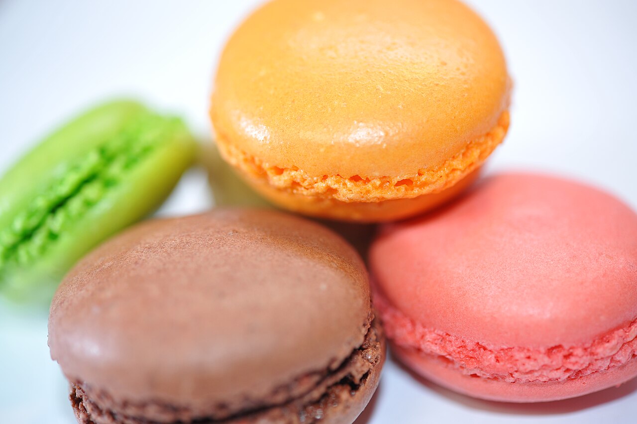 Four French macarons in orange, chocolate brown, pink, and lime green on a white surface