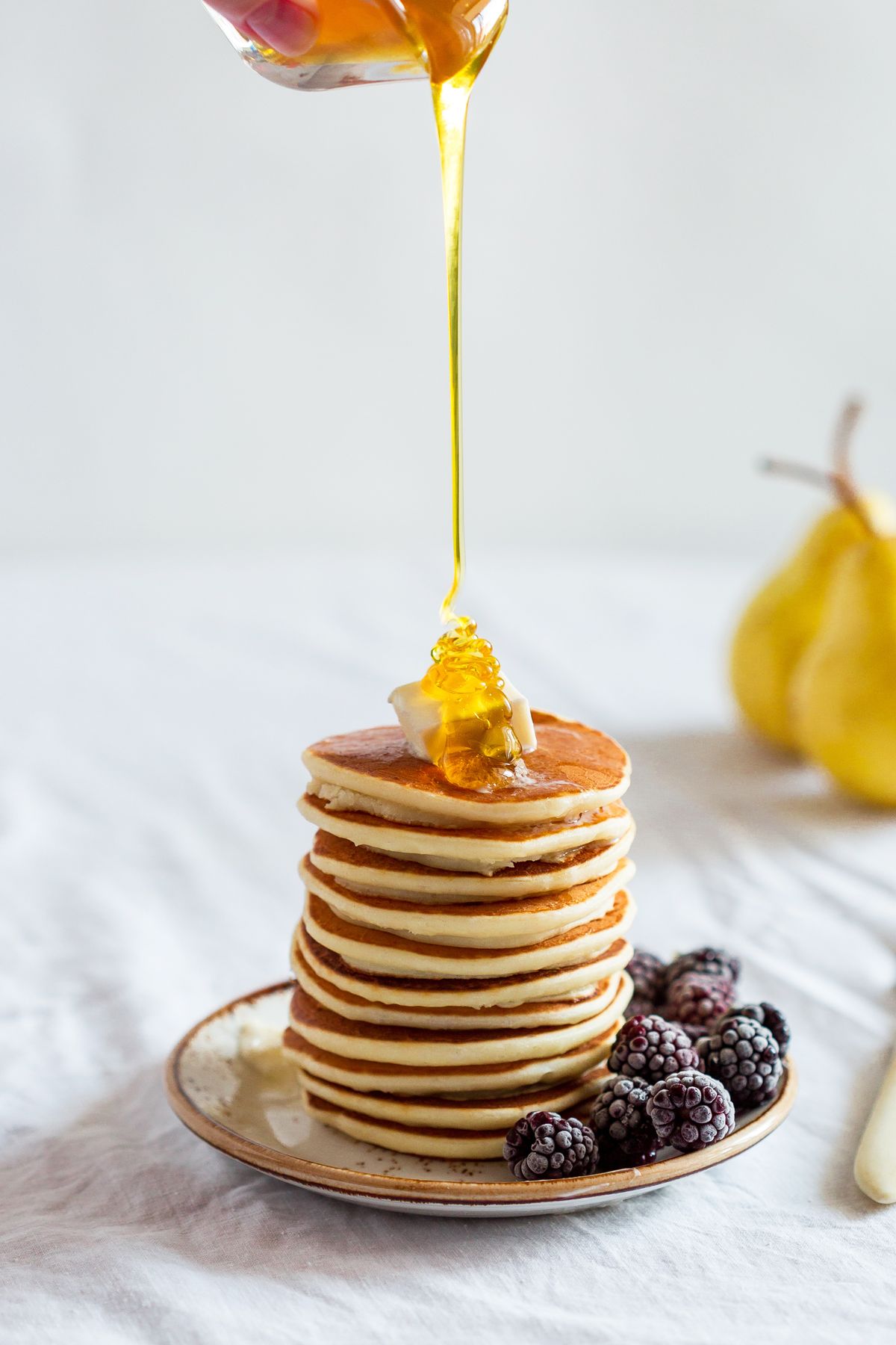 Tall stack of golden pancakes with butter as honey is poured, blackberries beside the plate